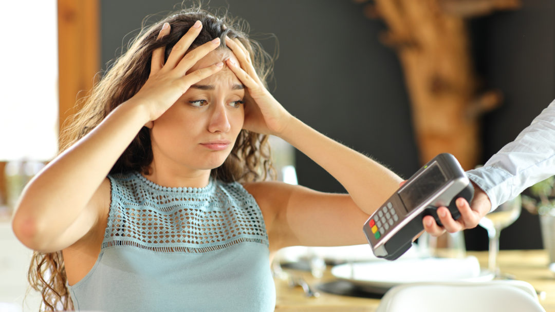 A worried person throws their hands on her head as a restaurant server extends a payment terminal toward them to pay the bill.