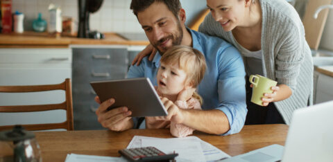 A happy family sitting at the kitchen table, reviewing financial documents on a tablet, with a laptop and calculator nearby.