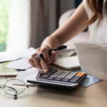 A woman calculating expenses at a desk, holding a receipt while using a calculator, with financial documents and a notebook in front of her.