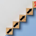 Wooden blocks arranged in a staircase pattern with black upward arrows, leading to a final block with a red percentage symbol, symbolizing financial growth.