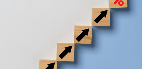 Wooden blocks arranged in a staircase pattern with black upward arrows, leading to a final block with a red percentage symbol, symbolizing financial growth.