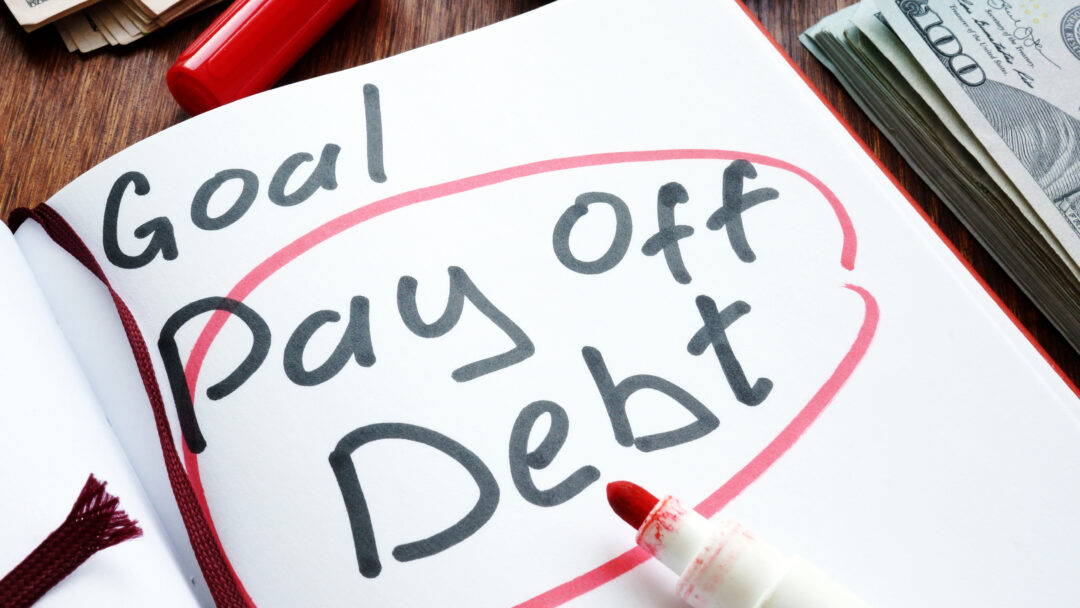 Notebook with "Goal: Pay Off Debt" written in bold letters, surrounded by a red circle, next to a red marker and stacks of cash.