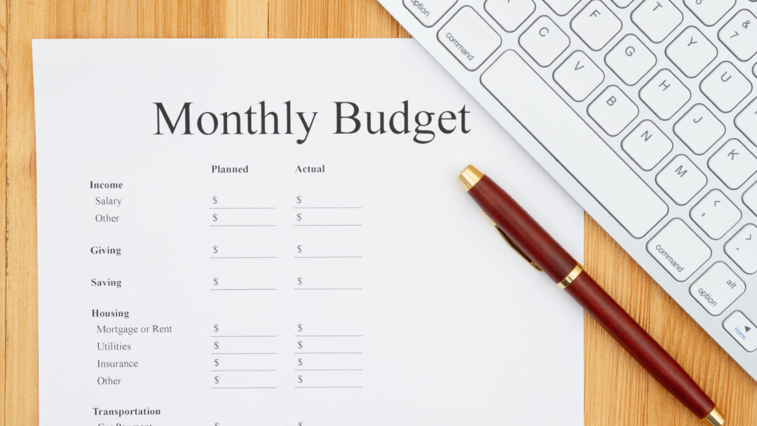 A "Monthly Budget" worksheet on a wooden desk, accompanied by a red and gold pen and a partial view of a white computer keyboard.