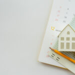 A calendar, miniature house model, calculator, and financial documents arranged on a desk, symbolizing mortgage planning and homeownership.